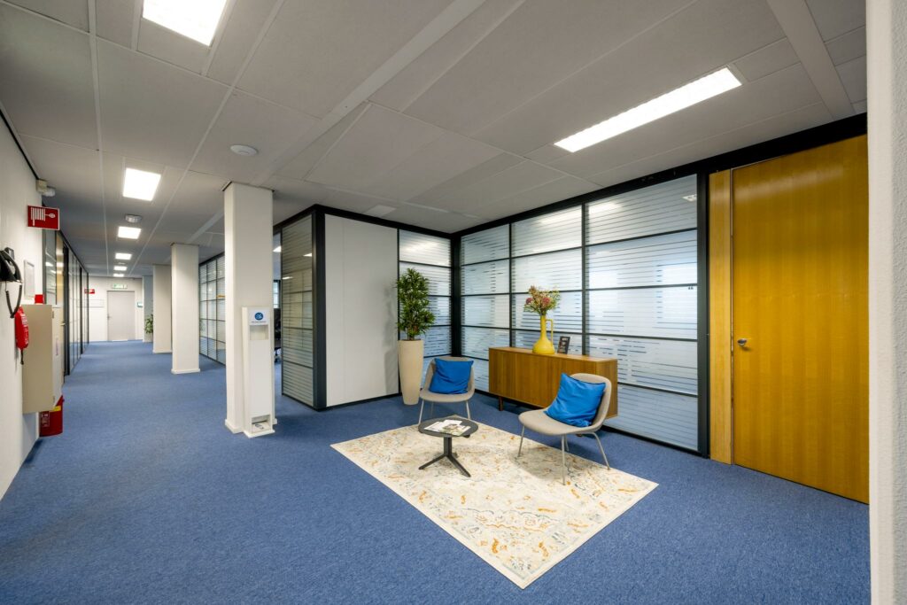 Moderne kantoorhal aan de Joop Geesinkweg met blauwe vloerbedekking, glazen wanden en een wachthoek met twee stoelen.